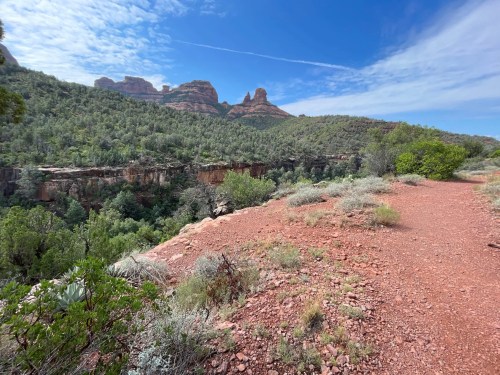A scenic view of a red dirt path winding through lush greenery, with rocky formations and a blue sky in the background.