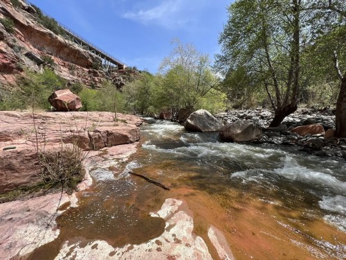 A serene river flows through a rocky landscape, surrounded by green trees and a clear blue sky.