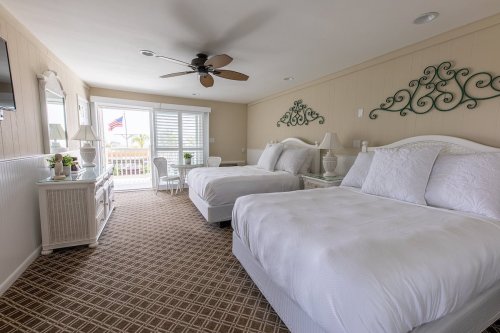 Bright hotel room with two beds, a ceiling fan, and a balcony view, featuring light decor and a cozy atmosphere.