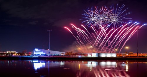 Colorful fireworks illuminate the night sky above a calm lake, reflecting vibrant lights from a nearby building.