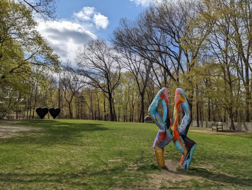 Colorful abstract sculpture in a grassy park with trees and two large heart-shaped sculptures in the background.