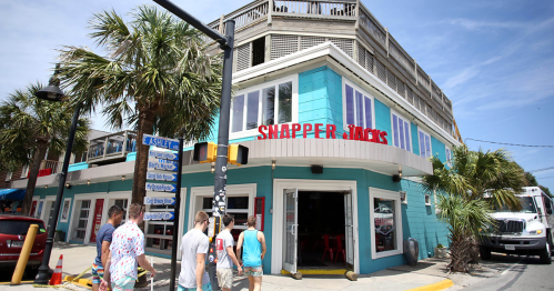 A colorful beachside restaurant named Snapper Jack's, with palm trees and people walking nearby.