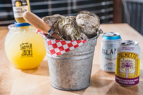 A metal bucket filled with oysters, a wooden shucker, and drinks including a cocktail and two beer cans.