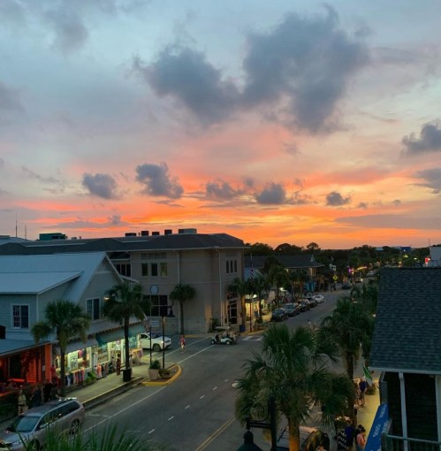 A vibrant sunset over a street lined with palm trees and shops, casting warm colors in the sky.