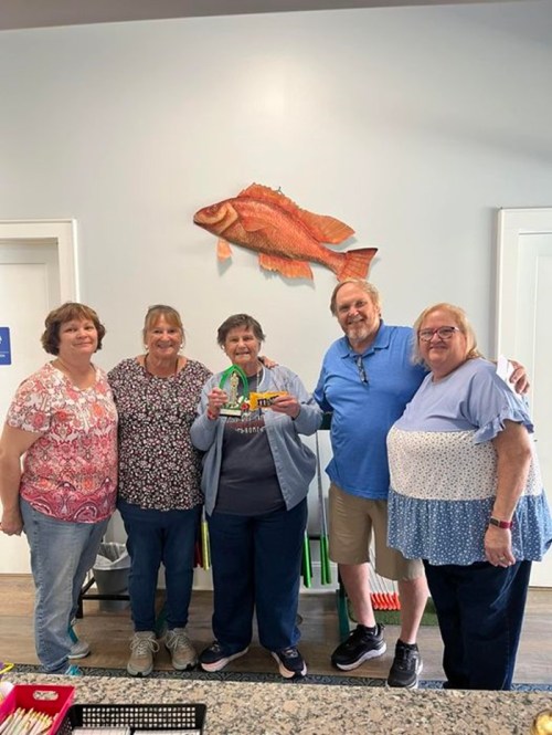 Five people stand together in a bright room, smiling, with a large fish decoration on the wall behind them.