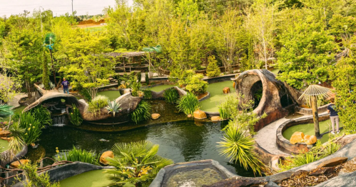 Lush garden with a pond, rock formations, and greenery, featuring pathways and small waterfalls.