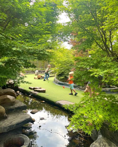 Two children playing mini-golf in a lush, green garden with rocks and a small pond.