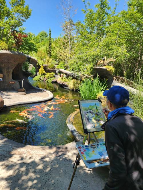 An artist paints by a serene pond filled with koi fish, surrounded by lush greenery and natural rock formations.