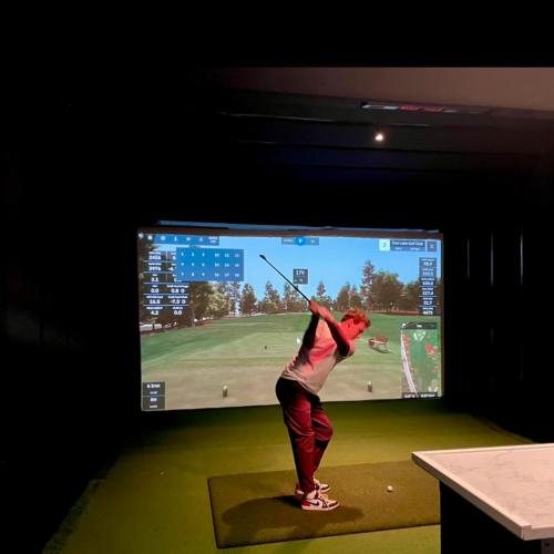 A person swings a golf club in front of a digital golf simulator displaying a course and stats.