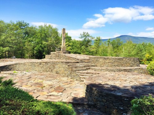 Stone steps lead to a tall wooden post, surrounded by greenery and mountains under a blue sky with clouds.