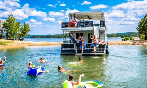A group of people swimming and playing in a lake near a houseboat on a sunny day with blue skies and trees in the background.