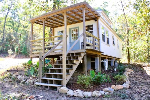 A small, elevated house with a porch and stairs, surrounded by greenery and rocks in a wooded area.