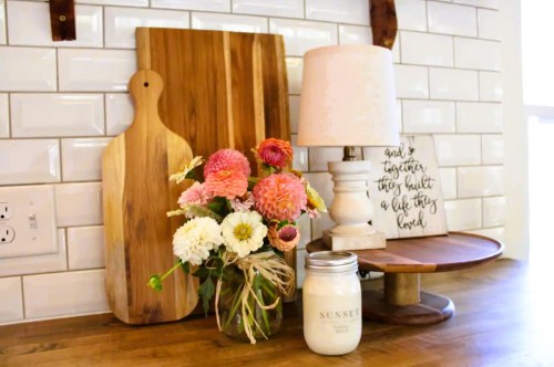 A cozy kitchen scene featuring a vase of flowers, a lamp, a cutting board, and a candle on a wooden countertop.