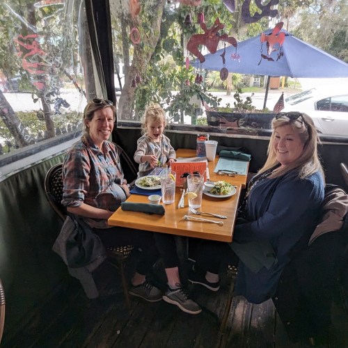 Three people sit at a table in a restaurant, enjoying meals and smiling, with greenery visible outside.