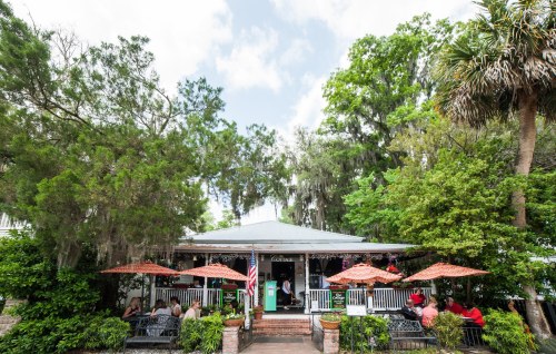A charming restaurant with outdoor seating, surrounded by lush greenery and trees, under a partly cloudy sky.