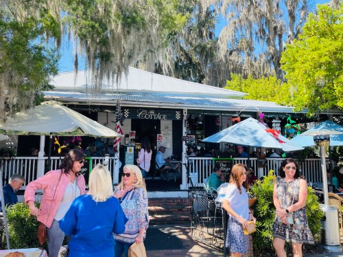 A charming restaurant with outdoor seating, surrounded by greenery and Spanish moss, on a sunny day.