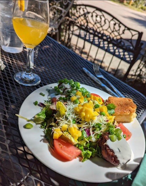 A colorful salad with mango, tomatoes, and cheese, served with cornbread and a glass of mimosa on a patio table.