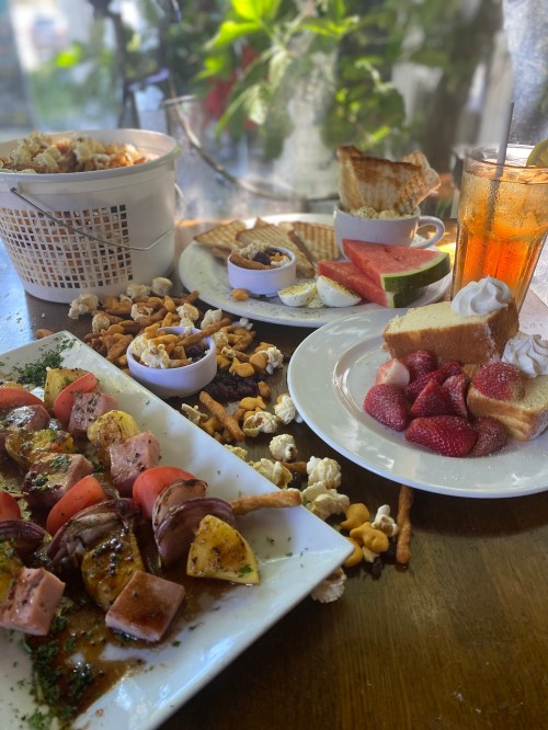 A table spread with skewers, fruit, popcorn, cake, and iced tea, set against a bright, leafy background.