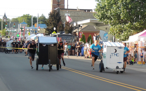 Participants race in quirky carts down a street lined with spectators and festival booths.