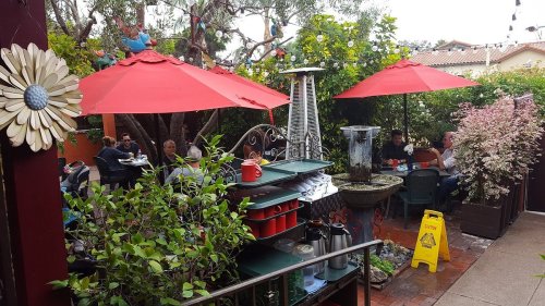 Outdoor dining area with red umbrellas, greenery, and a fountain, featuring patrons enjoying their meals.