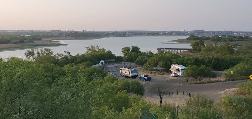 A scenic view of a lake surrounded by greenery, with RVs parked near the water's edge and a distant town visible.