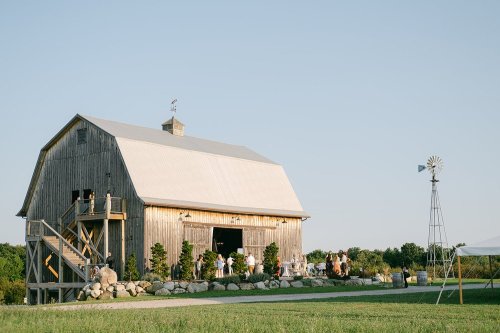 A rustic barn venue with guests mingling outside, surrounded by greenery and a windmill in the background.