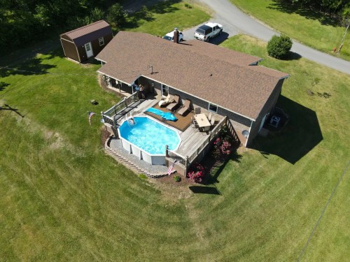 Aerial view of a house with a pool, surrounded by a deck, lawn, and trees, with a shed and parked vehicle nearby.