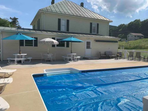 A backyard pool area with a blue cover, patio furniture, umbrellas, and a house in the background on a sunny day.