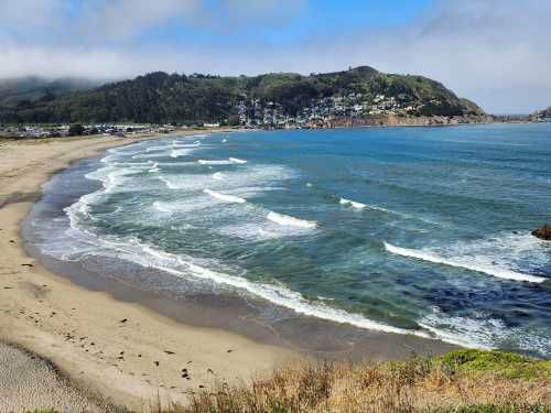 A scenic view of a beach with gentle waves, surrounded by green hills and a small coastal town in the background.