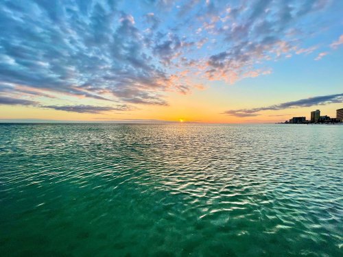 A serene sunset over calm waters, with colorful clouds reflecting in the sky and a distant shoreline.