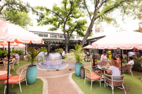 Outdoor dining area with colorful umbrellas, a fountain, and people enjoying meals in a lush, green setting.