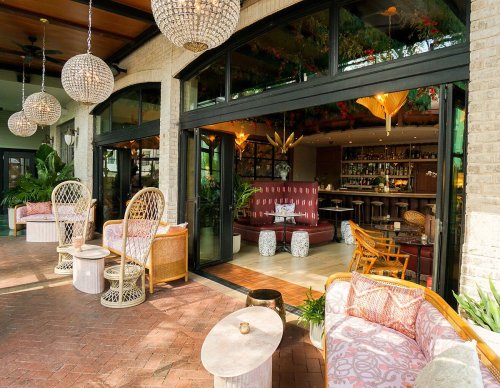 A stylish lounge with rattan chairs, elegant lighting, and a bar, surrounded by greenery and large glass doors.