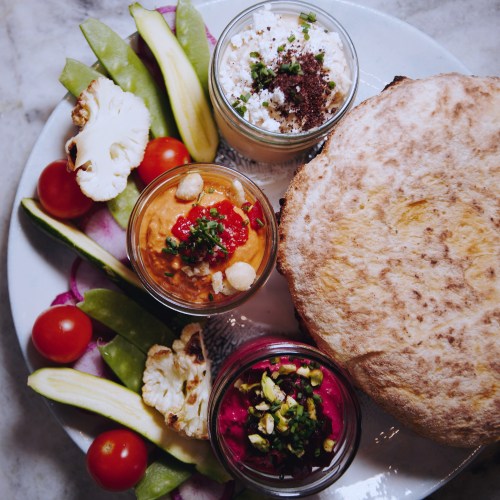 A platter featuring dips in jars, fresh vegetables, and a flatbread, arranged artfully on a marble surface.