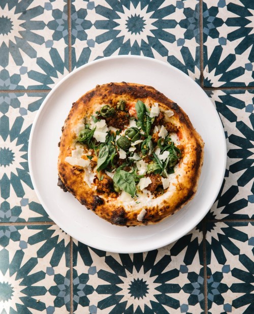 A freshly baked pizza topped with greens and cheese, served on a white plate against a patterned blue tile background.