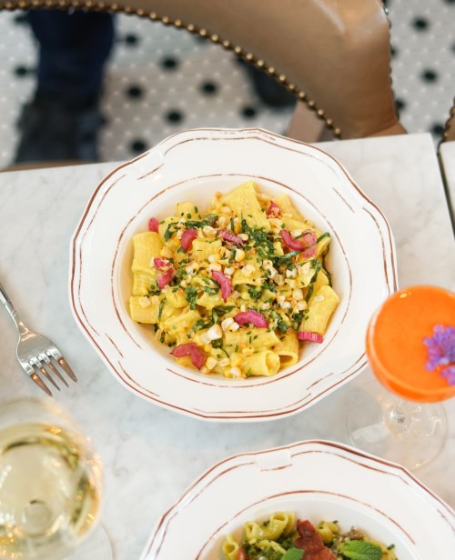 A bowl of pasta with corn, greens, and pink radish, accompanied by a cocktail and a glass of white wine.