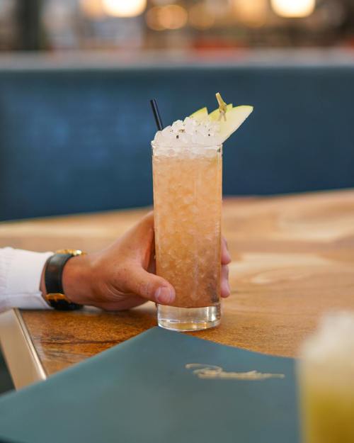A hand holding a tall glass of a refreshing cocktail with ice and a slice of fruit, placed on a wooden table.