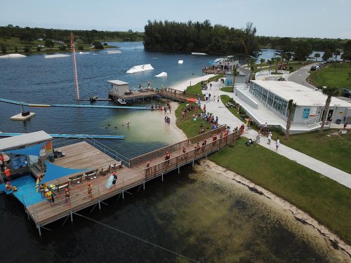 Aerial view of a lakeside park with people enjoying water activities, a dock, and a modern building nearby.