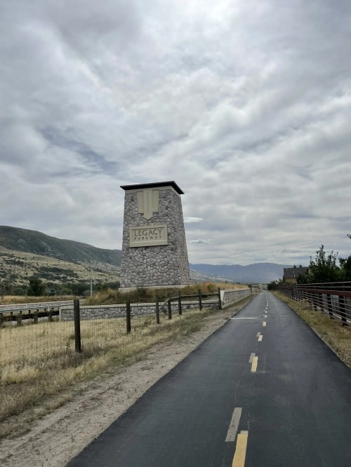 A stone monument with "Legacy" engraved, beside a paved path and grassy fields under a cloudy sky.