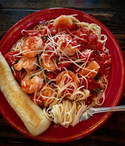 A red plate with shrimp pasta in tomato sauce, topped with cheese, and a piece of bread on the side.
