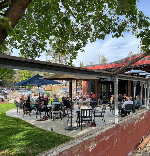 A lively outdoor dining area with people seated at tables under umbrellas, surrounded by greenery and a red building.