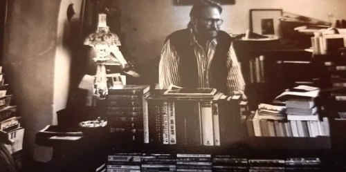 A black-and-white photo of a man in a vest surrounded by stacks of books in a cozy, cluttered room.