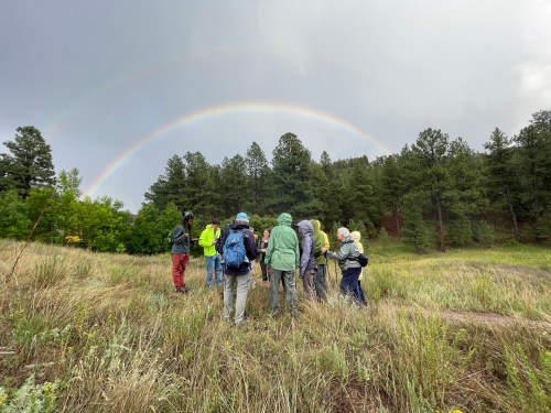 A group of people in rain gear stands in a grassy field with a rainbow arching over a forested background.