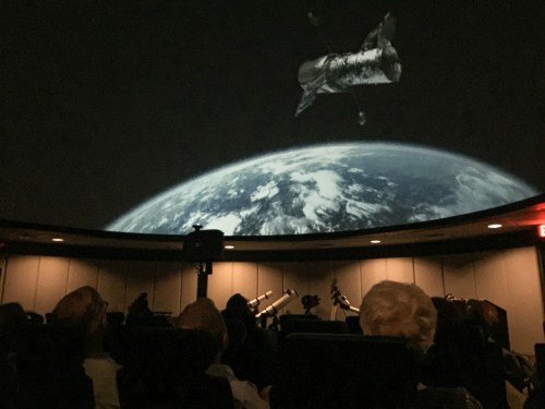 A planetarium scene showing a spacecraft above Earth, with an audience seated in the foreground.