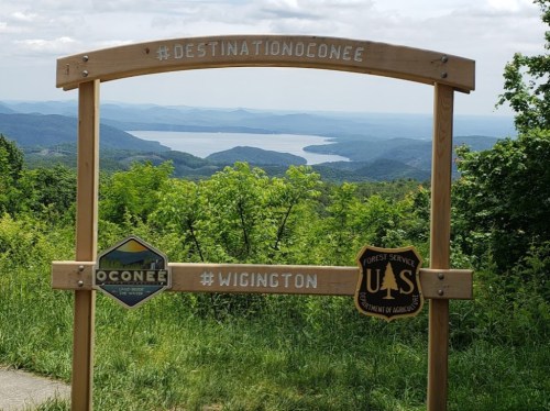 A wooden sign with hashtags and badges overlooks a scenic view of mountains and a lake in the distance.