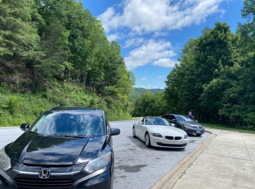 Three cars parked along a scenic road, surrounded by lush green trees and a blue sky with fluffy clouds.