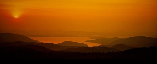 A serene sunset over layered mountains and a calm lake, casting warm orange and yellow hues across the sky.