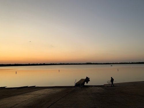 A serene lakeside at sunset, with a lone figure near the water's edge and gentle reflections on the surface.