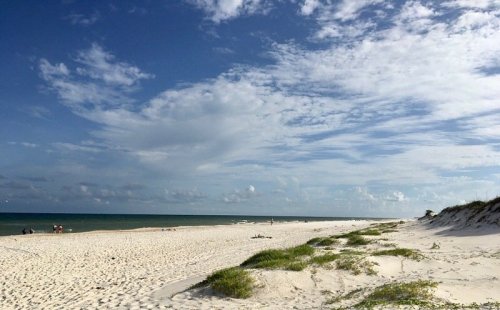A serene beach scene with soft white sand, gentle waves, and a blue sky dotted with clouds.