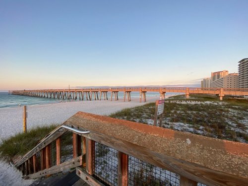A wooden pier extends over a sandy beach, with tall buildings in the background and a clear sky at sunset.