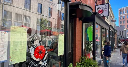 A storefront of Early Girl Eatery featuring a tomato logo and menus displayed in the window, with people walking by.